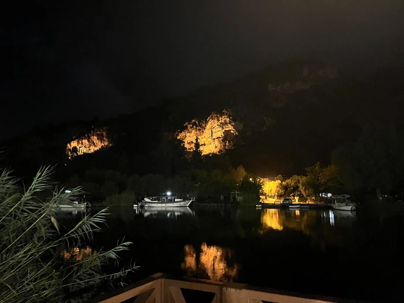 The Kaunos rock tombs floodlit at night above the river, boats moored below The Kaunos rock tombs floodlit at night above the river, boats moored below