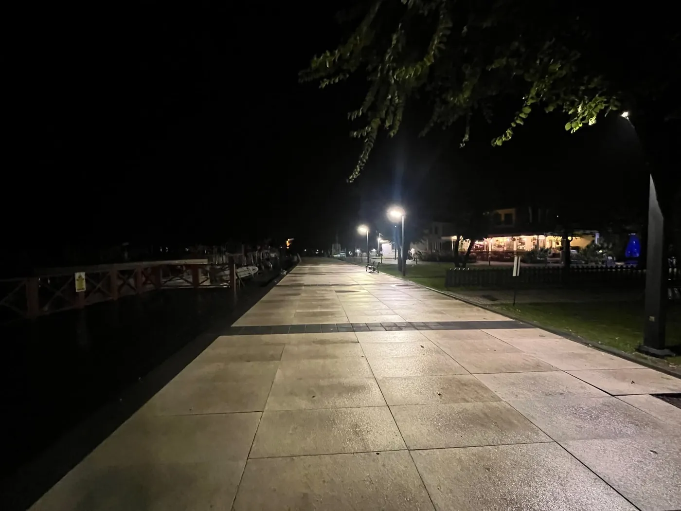 Die gepflasterte Promenade bei Nacht, voraus ein beleuchtetes Café, links liegen Boote
